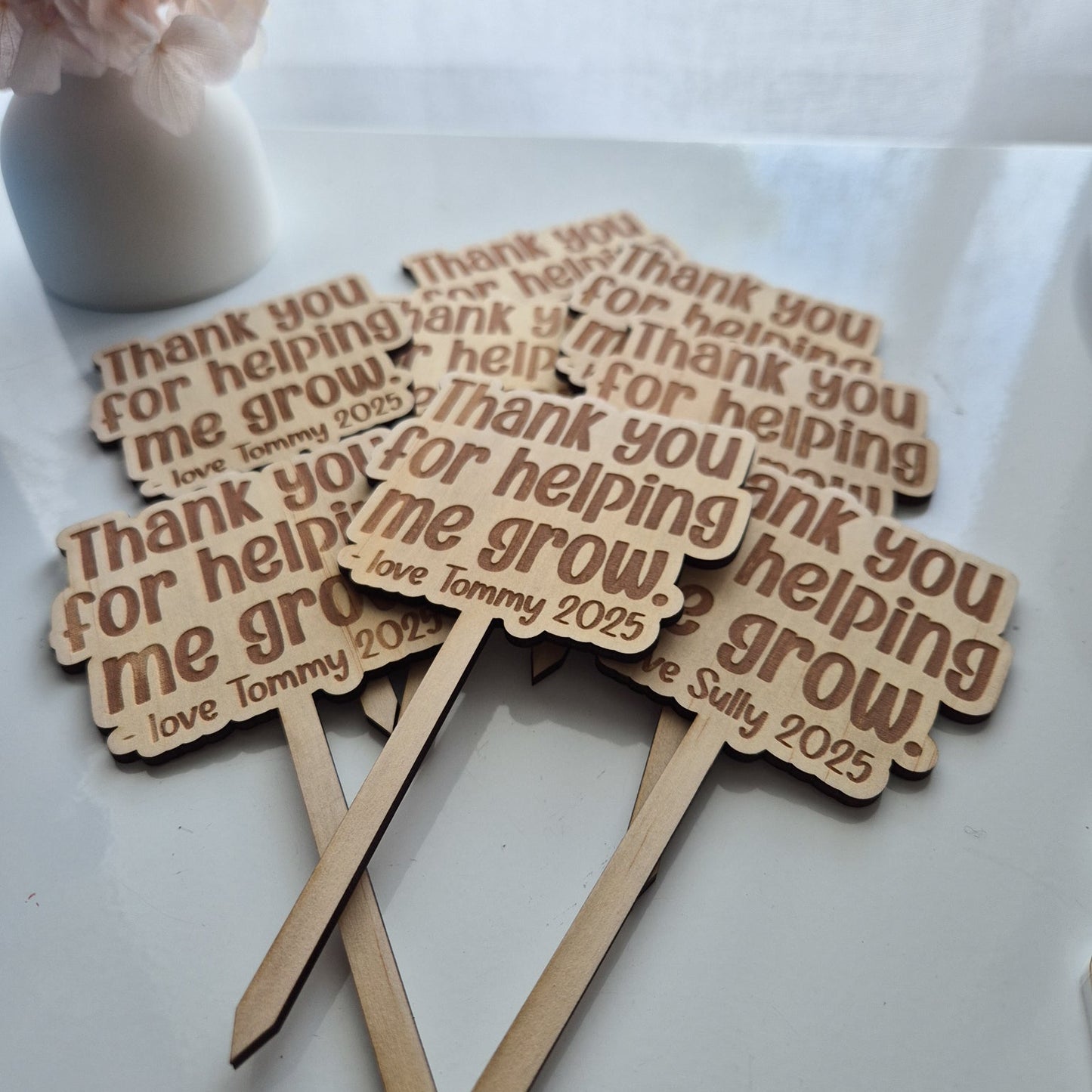 Wooden plant stakes with engraved text on a white surface with flowers in the background.