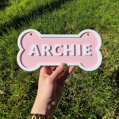 Pink bone-shaped sign with 'ARCHIE' on it held by a hand against a grassy background