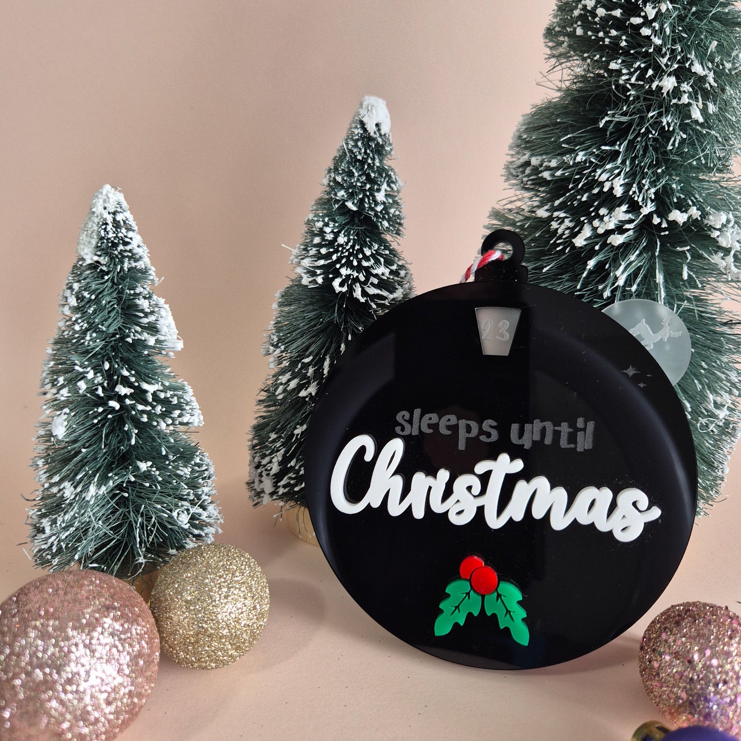 Black ornament with 'Sleeps until Christmas' text, surrounded by Christmas trees and ornaments on a beige background